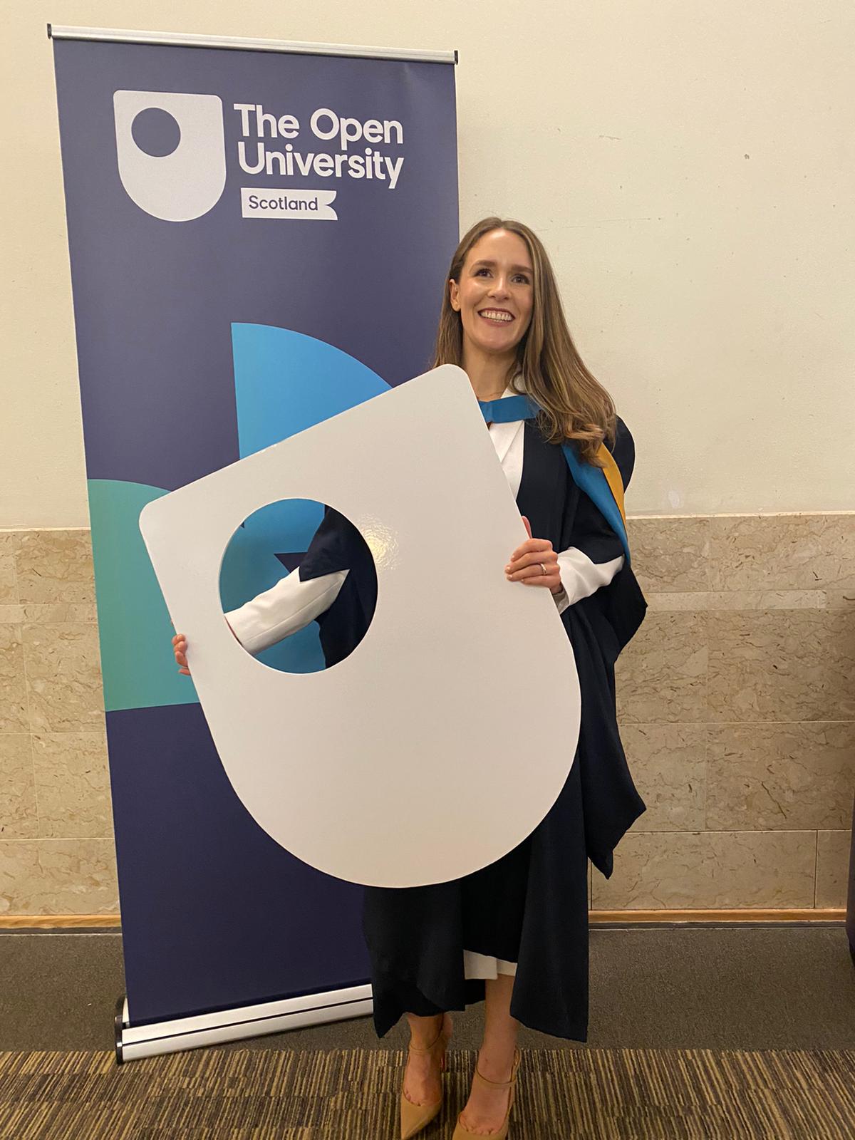 Alex at her graduation standing in front of an Open University banner and holding a large cardoboard cutout of the 
                        Open Univeristy logo